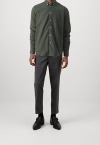 Mężczyzna w ciemnozielonej koszuli zapinanej na guziki, szarych dopasowanych spodniach, czarnych skarpetach i czarnych skórzanych butach eleganckich, stojący na jednolitym jasnym tle.