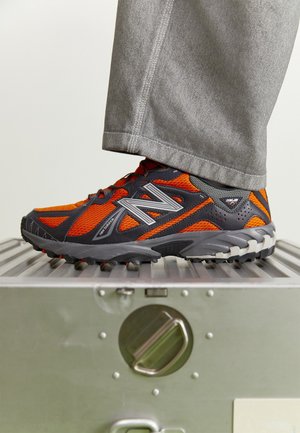 Henkilö, jolla on harmaat housut ja oranssi-musta New Balancen trail-juoksukengät, astuu hopeanvärisen metallilaatikon päälle.