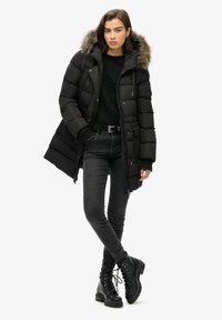 Abrigo negro tipo puffer con capucha de pelo, frente con cremallera y textura de punto por debajo. Lleva unos jeans skinnies negros y botas de cordones.