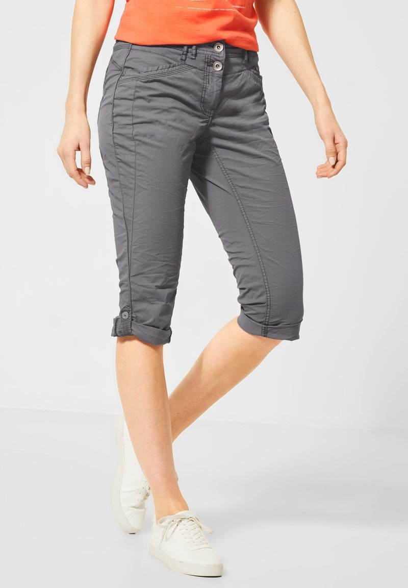 Femme portant un pantalon capri gris retroussé, un haut orange et des baskets blanches, debout avec une jambe légèrement pliée.