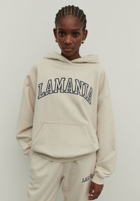 Beige kapucnis pulóver "LAMANIA" grafikai mintával sötétkék színben, elülső zsebbel, húzózsinórral ellátott kapucnival és bordás mandzsettákkal. A megfelelő bézs nadrág is látható.