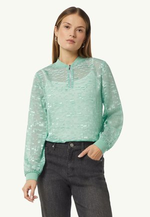 Mintgrünes, durchsichtiges Blusentop mit dezentem Texturmuster, Reißverschluss-Ausschnitt und elastischen Bündchen, kombiniert mit dunklen Jeans.