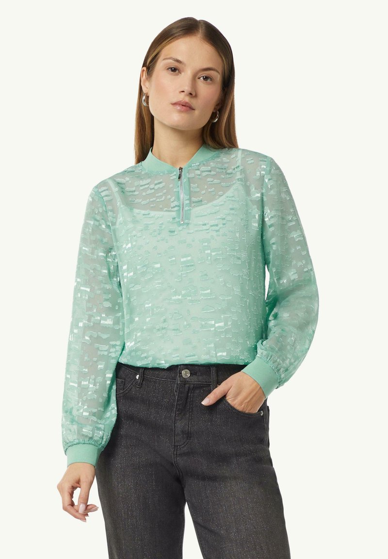 Mintgrünes, durchsichtiges Blusentop mit dezentem Texturmuster, Reißverschluss-Ausschnitt und elastischen Bündchen, kombiniert mit dunklen Jeans.