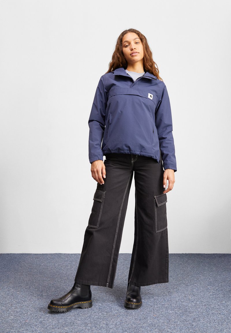 Marineblaues Pullover-Jacke mit Frontreißverschluss und Brusttasche; kombiniert mit weiten schwarzen Cargo-Hosen, die seitliche Taschen und weiße Nähte haben. Schwarze Knöchelboots.