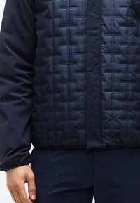 Άνδρας που φοράει ναυτικό μπλε μπουφάν με υφασμένο διαγώνιο μοτίβο και αντίστοιχο ναυτικό μπλε παντελόνι, με το χέρι χαλαρά στο πλάι.