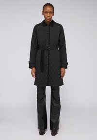 Černý prošívaný kabát s límcem, zapínáním na knoflíky, detailem pásku v oblasti pasu a dlouhými rukávy. Nositý přes černé kožené kalhoty a kotníkové boty.