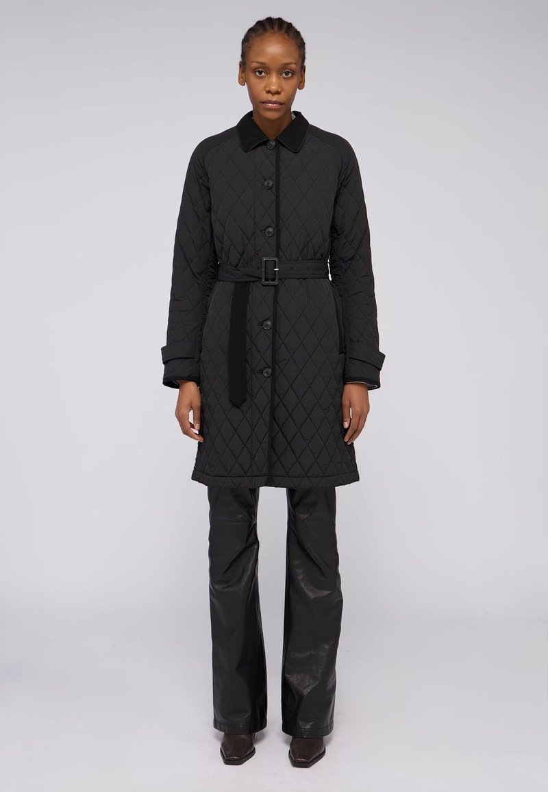 Černý prošívaný kabát s límcem, zapínáním na knoflíky, detailem pásku v oblasti pasu a dlouhými rukávy. Nositý přes černé kožené kalhoty a kotníkové boty.