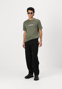 Olive green T-shirt with a logo, paired with black pants and black shoes. Accessories include dark sunglasses. Minimalist, casual design.