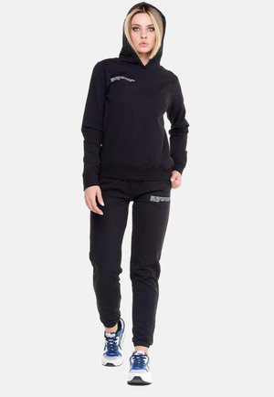 Une femme porte un sweat-shirt noir à capuche et un pantalon de jogging assorti, avec des détails de texte blancs et des baskets bleues et blanches.