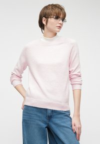 Maglione rosa chiaro con finitura testurizzata, polsini e orlo a coste, e scollo rotondo, abbinato a jeans a gamba larga blu.
