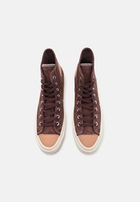 Høye sneakers i mørkebrunt semsket skinn med beige tåhette, hvit gummisåle og brune skolisser. Har sydd detaljer og metalløyler.