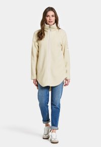 Cremefarvet fleecejakke med høj krave, fuld frontlynlås og sidelommer; parret med blå jeans og grå sneakers.
