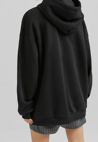 Ylisuuri musta huppari, pehmeällä pinnalla ja kiristysnauhalla varustettu huppu. Varustettu lipsahtaneilla harteilla ja resorilla varustetulla helmalla, yhdistetty raidoitettuihin shortseihin.