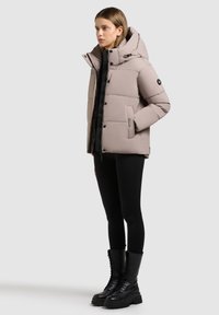 Hellbeige wattierte Jacke mit hohem Kragen, Reißverschluss und Druckknöpfen, schwarzen Akzenten und Taschen, kombiniert mit schwarzen Leggings und Schnürstiefeln.