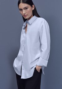 Vaaleanharmaa pitkähihainen nappipaita, jossa on terävä kaulus ja rento istuvuus, yhdistetty mustiin housuihin. Sileä, minimalistinen muotoilu.