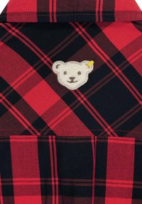 Camicia a quadri rossa e nera con una superficie morbida e testurizzata. Presenta una toppa ricamata a forma di orso bianco con accenti gialli.