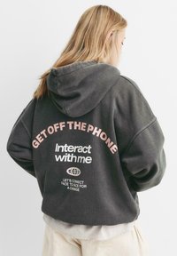Grauer Hoodie mit lockerer Passform und Kapuze. Auf der Rückseite mit pinkem Text: "Geh vom Telefon weg" und "Interagiere mit mir", mit zusätzlichem Text darunter.