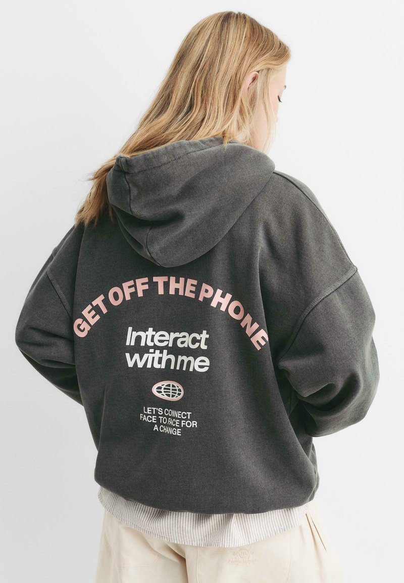 Grauer Hoodie mit lockerer Passform und Kapuze. Auf der Rückseite mit pinkem Text: "Geh vom Telefon weg" und "Interagiere mit mir", mit zusätzlichem Text darunter.