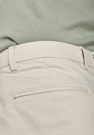 Primo piano di pantaloni beige dalla texture pronunciata con tasca posteriore a filetto e passanti per cintura, abbinati a un top in tessuto verde chiaro.