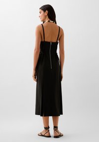 Schwarzes Midi-Kleid mit dünnen Trägern, Reißverschluss im Rücken und seitlichen Bindebändern. Dazu minimalistische schwarze Sandalen mit gewickelten Riemen. Glattes Material.