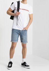 Biała koszulka z czerwonym napisem "ILL ROMANCE", jasnoniebieskie szorty dżinsowe, czarne sneakersy i białe skarpetki do kostek. Ciemny plecak zawieszony na ramieniu.