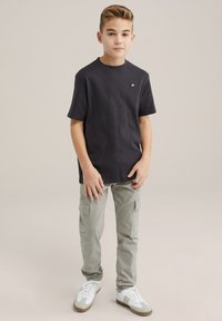 T-shirt nera in cotone con maniche corte e piccolo logo, abbinata a pantaloni cargo grigi chiari con tasche laterali e sneakers bianche.