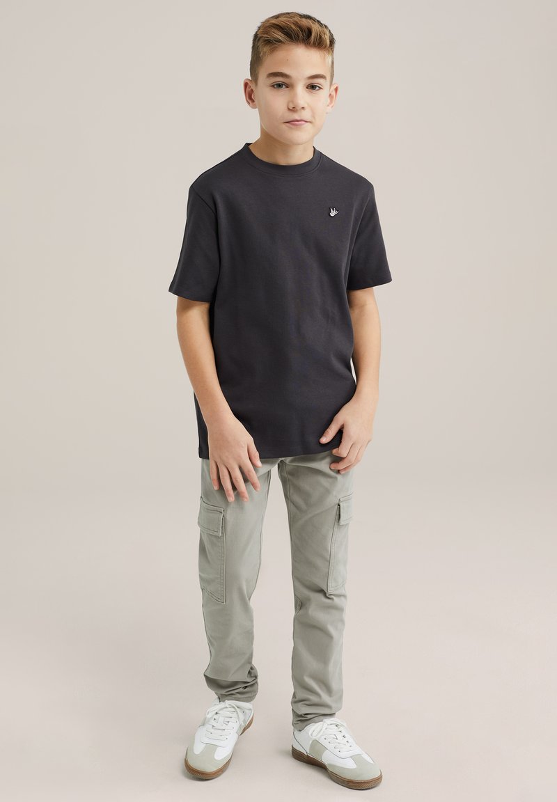 T-shirt nera in cotone con maniche corte e piccolo logo, abbinata a pantaloni cargo grigi chiari con tasche laterali e sneakers bianche.