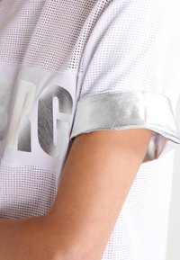 Bílý síťovaný T-shirt s metalickými stříbrnými manžetami a výrazným stříbrným nápisem "BRG" na přední straně, zdůrazňující prodyšný design.