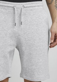Shorts en coton gris avec cordon de serrage, présentant des éléments en métal. Le tissu a une texture subtile, avec des poches latérales et une coupe décontractée.