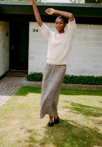 Hellbeige Strickpullover, maxi Rock mit schwarz-weißer Wellenmusterung, schwarze Mules, stehend auf Gras mit modernem Haus im Hintergrund.