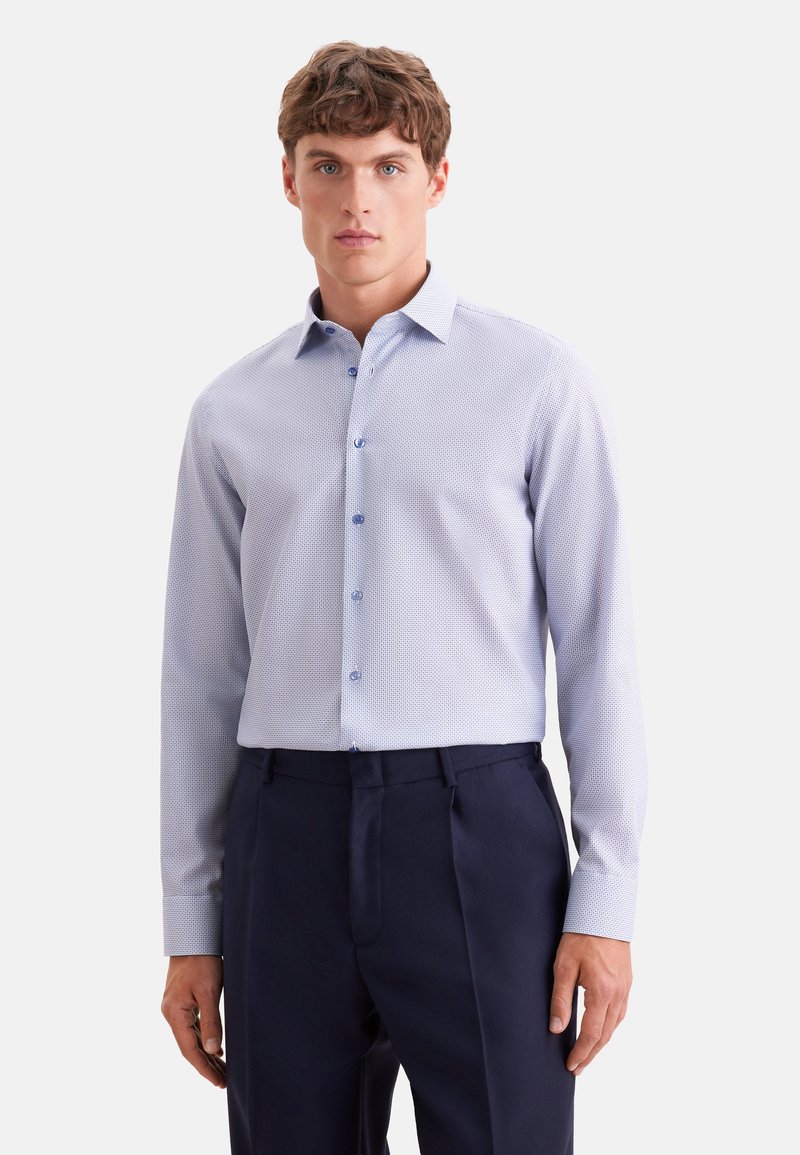 Jeune homme portant une chemise à manches longues bleu clair à motifs et un pantalon bleu foncé, debout devant un fond uni.