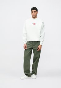 Bílá mikina s logem "TOMMY", zelené volné kalhoty a bílé tenisky. Mikina má kulatý výstřih a spadlá ramena.