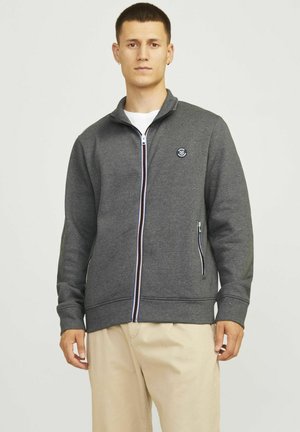 Grå zip-up sweatshirt med sidelommer, hvid inderside, ribbede manchetter og et logo patch på brystet. Glat stofstruktur.