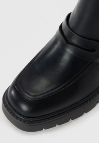 Bottine en cuir noir avec un bout arrondi, détail de sangle décorative et semelle en caoutchouc texturée avec un accent cousu autour du bord.