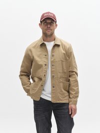 Veste en toile beige avec fermeture à boutons et deux poches avant, portée sur un t-shirt blanc, associée à un jean foncé et une casquette bordeaux.