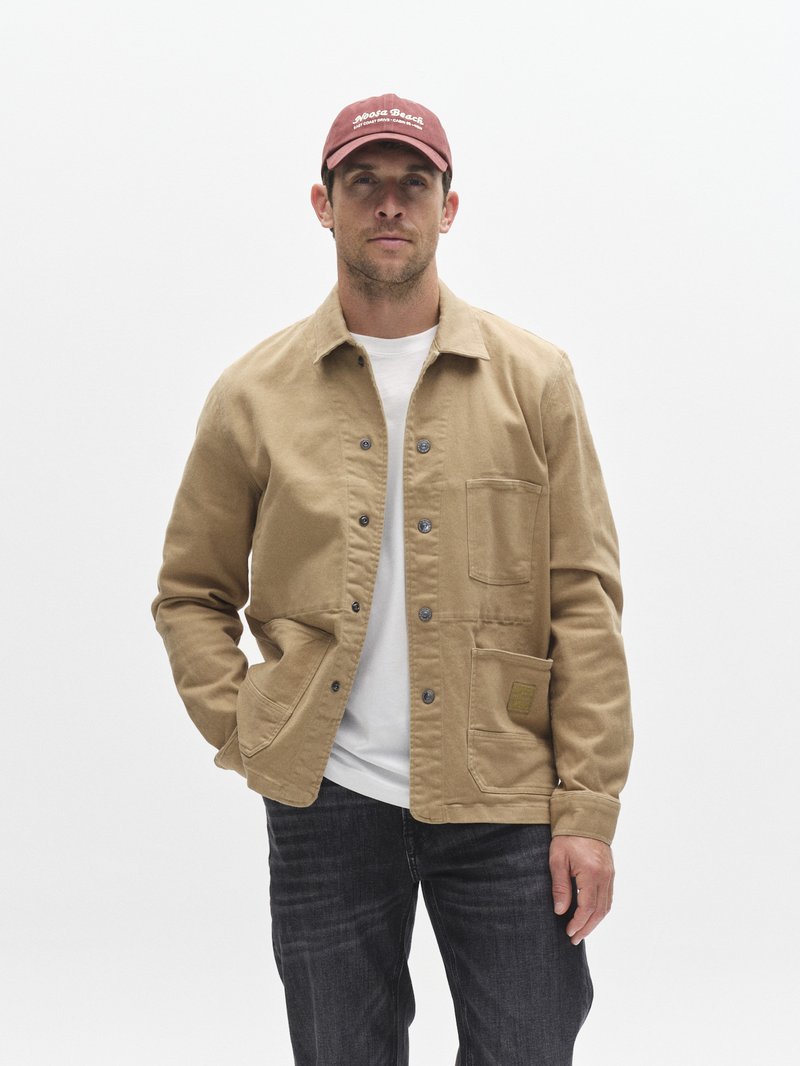 Veste en toile beige avec fermeture à boutons et deux poches avant, portée sur un t-shirt blanc, associée à un jean foncé et une casquette bordeaux.