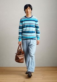 Pull bleu rayé, jean en denim clair et sac en cuir marron foncé. Fond de couleur naturelle avec un sol en bois. Tenue décontractée.