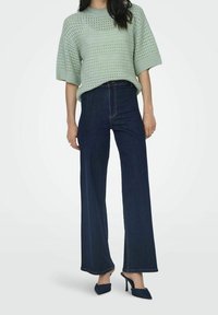 Ljusgrön stickad kortärmad tröja i kombination med högmidjade, vida mörkblå jeans. Har spetsiga marinblå klackar.