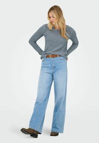 Femme blonde portant un pull gris en maille, un jean large taille haute bleu clair, une ceinture marron et des bottines marron, debout les mains sur les hanches.