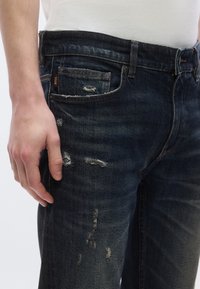 Sötétkék, vékony szárú farmernadrág, látható kopásokkal, enyhe bolyhosodással és elülső zsebbel. Gombos és cipzáras záródás.