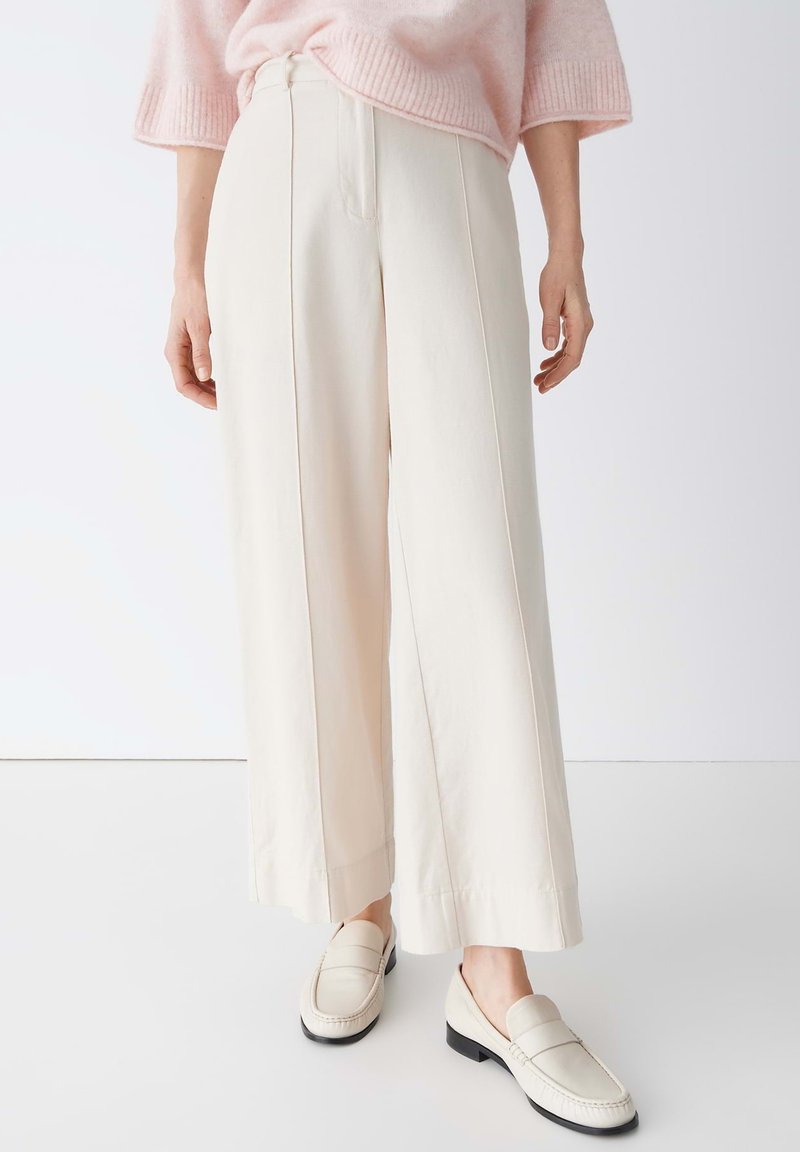Femme portant un pantalon large à taille haute couleur crème, associé à des mocassins crème assortis et un pull rose pâle.