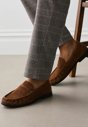 Rudos zomšos mokasinai dėvimi su pilkais languotais kelnėmis, viena koja ant grindų, o kita remiasi į medinę kėdės koją.