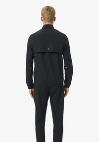 Man die met zijn rug naar de camera staat, gekleed in een zwarte jas met een elastische tailleband en bijpassende zwarte broek, tegen een effen witte achtergrond.