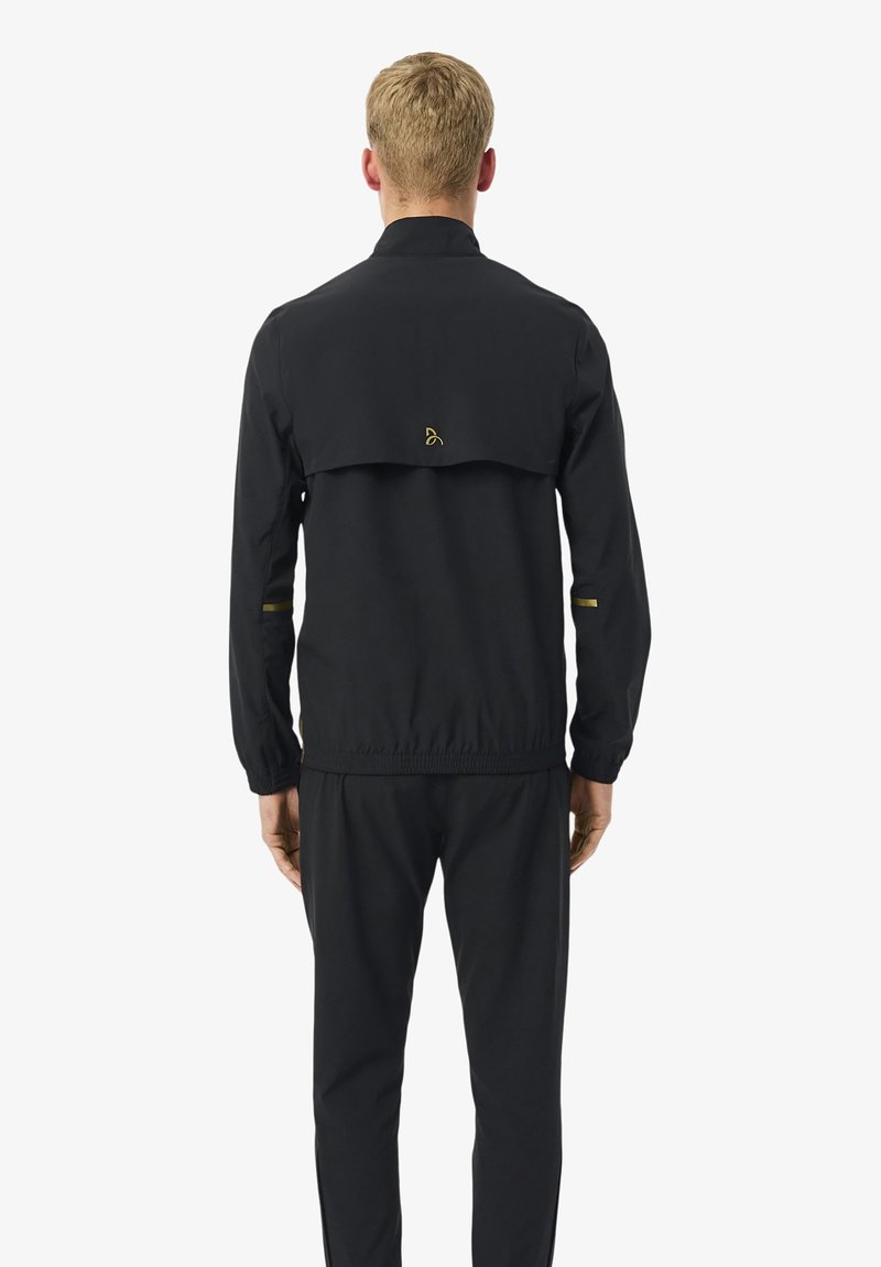 Mann, der von hinten steht, trägt eine schwarze Jacke mit elastischem Bund und passende schwarze Hose vor einem schlichten weißen Hintergrund.