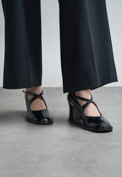 Black patent leather heels with a rounded toe and prominent block heel, featuring crisscross straps and a cutout design.