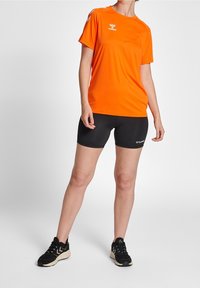 Jasnopomarańczowa koszulka sportowa z czarnym logo i akcentami, w połączeniu z czarnymi szortami i czarnymi butami sportowymi. Odpowiednia do aktywności sportowych.