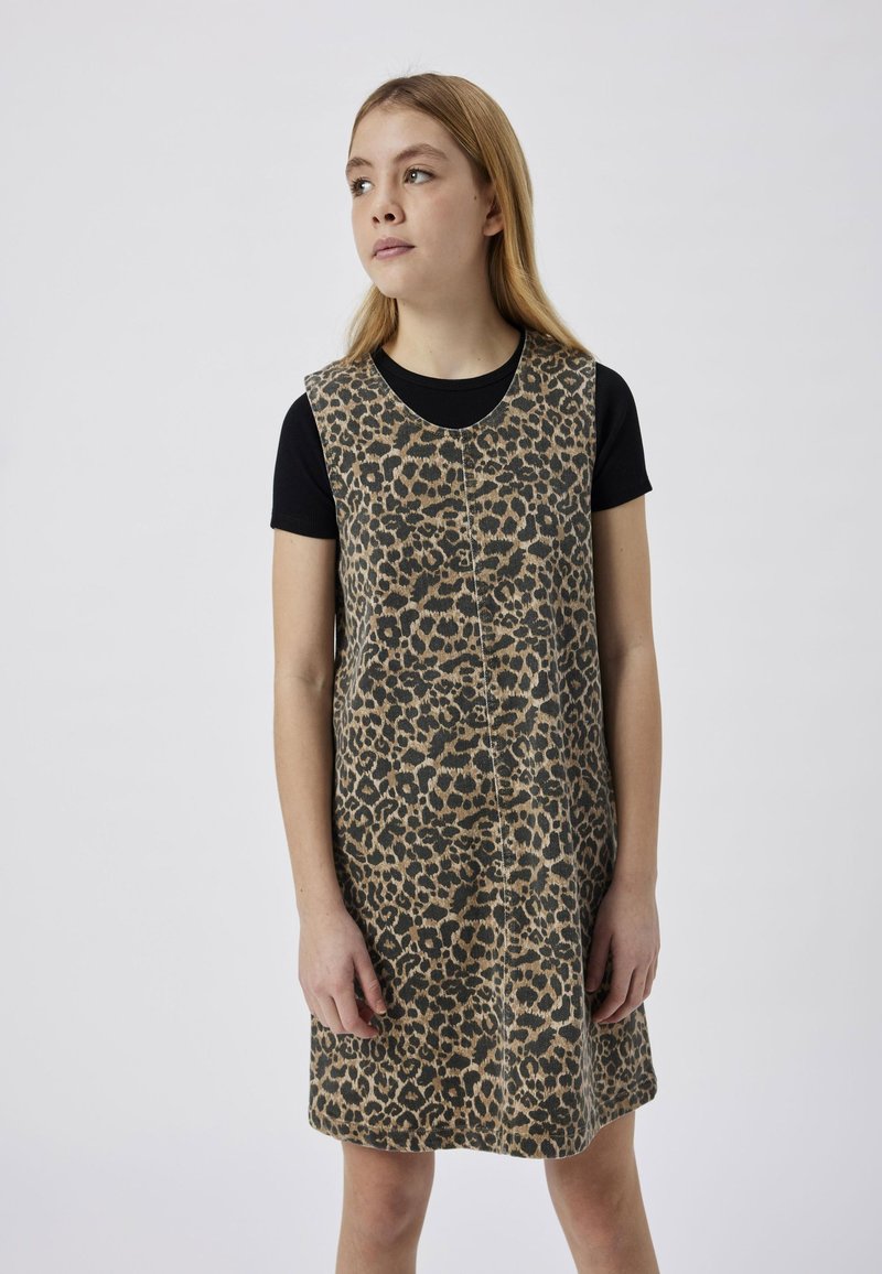 Giovane donna con capelli lisci castano chiaro che indossa un vestito senza maniche con stampa leopardata sopra una maglietta nera a maniche corte, guardando di lato.