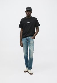 Black cotton t-shirt with "CHALÉT" text, paired with light blue denim jeans. Mid-rise fit, slight fade. White sneakers complete the look.