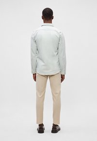 Ανοιχτό πράσινο πουκάμισο με μακριά μανίκια, συνδυασμένο με μπεζ παντελόνι και σκούρα καφέ παπούτσια, σε προοπτική από πίσω.