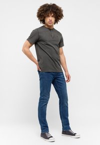 Tmavě šedé henley tričko s krátkým rukávem a hnědou knoflíkovou légou, v kombinaci s modrými slim-fit džíny a šedými teniskami.